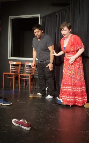 Two actors on a stage looking with surprise at two shoes sitting on the stage